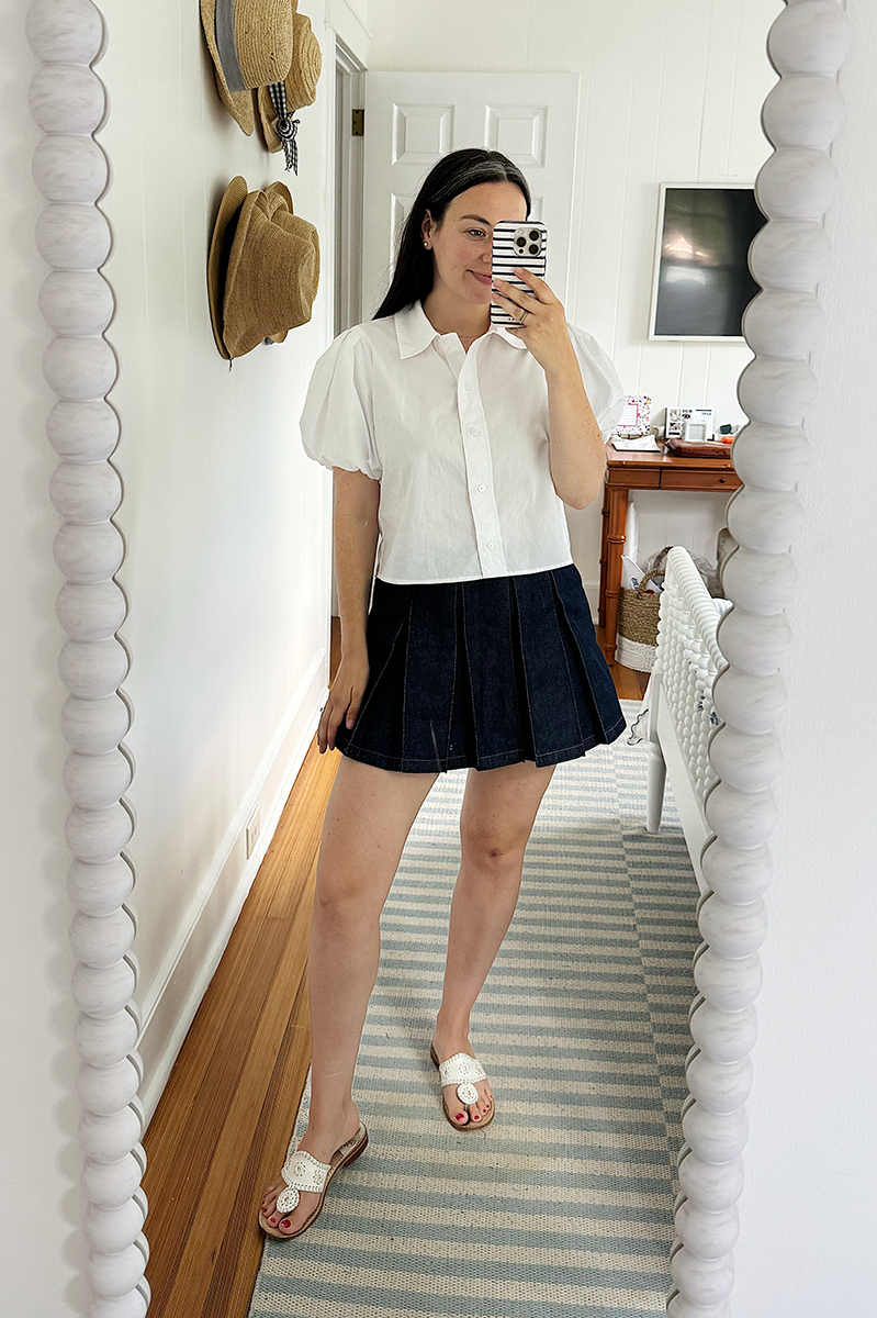 A mirror selfie of Carly wearing a white puff sleeve shirt with a dark denim pleated denim skort.