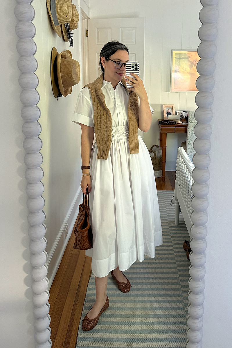 Carly taking a mirror selfie wearing a white dress and a camel sweater over her shoulders.