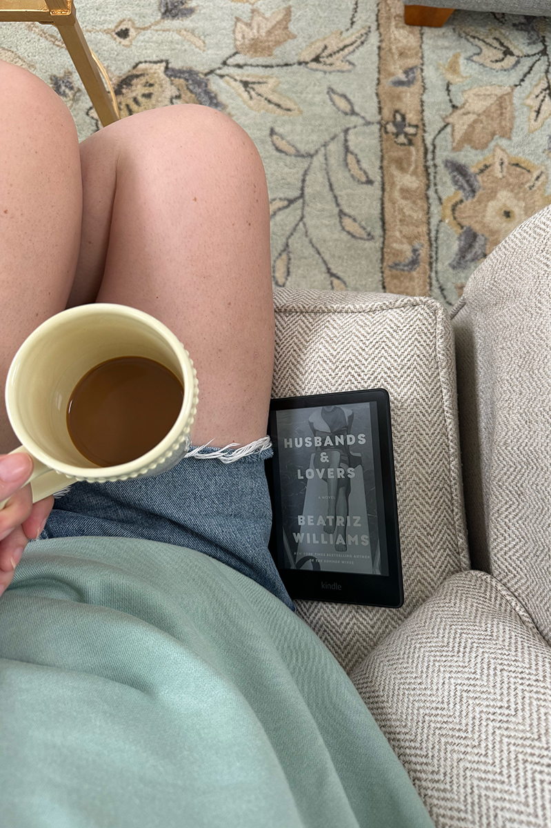 Carly wearing denim shorts and a sweatshirt with her kindle and coffee cup. 