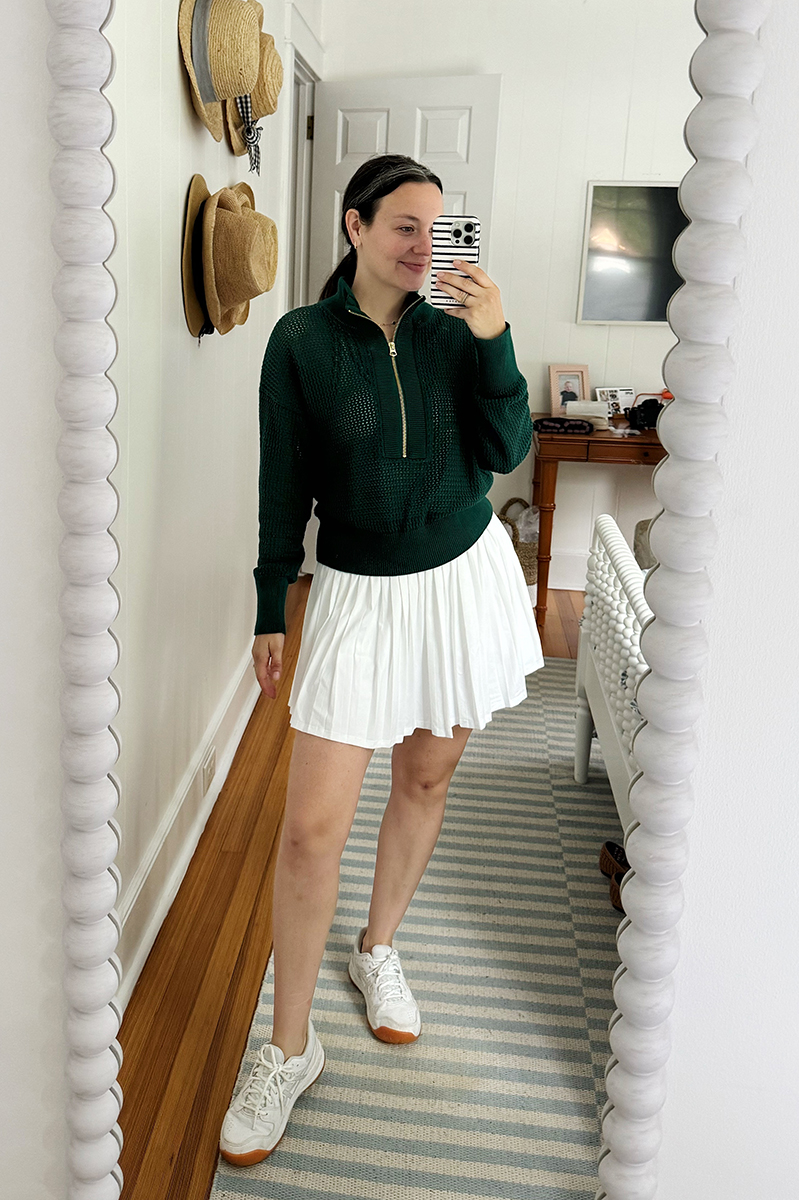 Carly taking a mirror selfie wearing a white tennis dress and a green sweatshirt over.