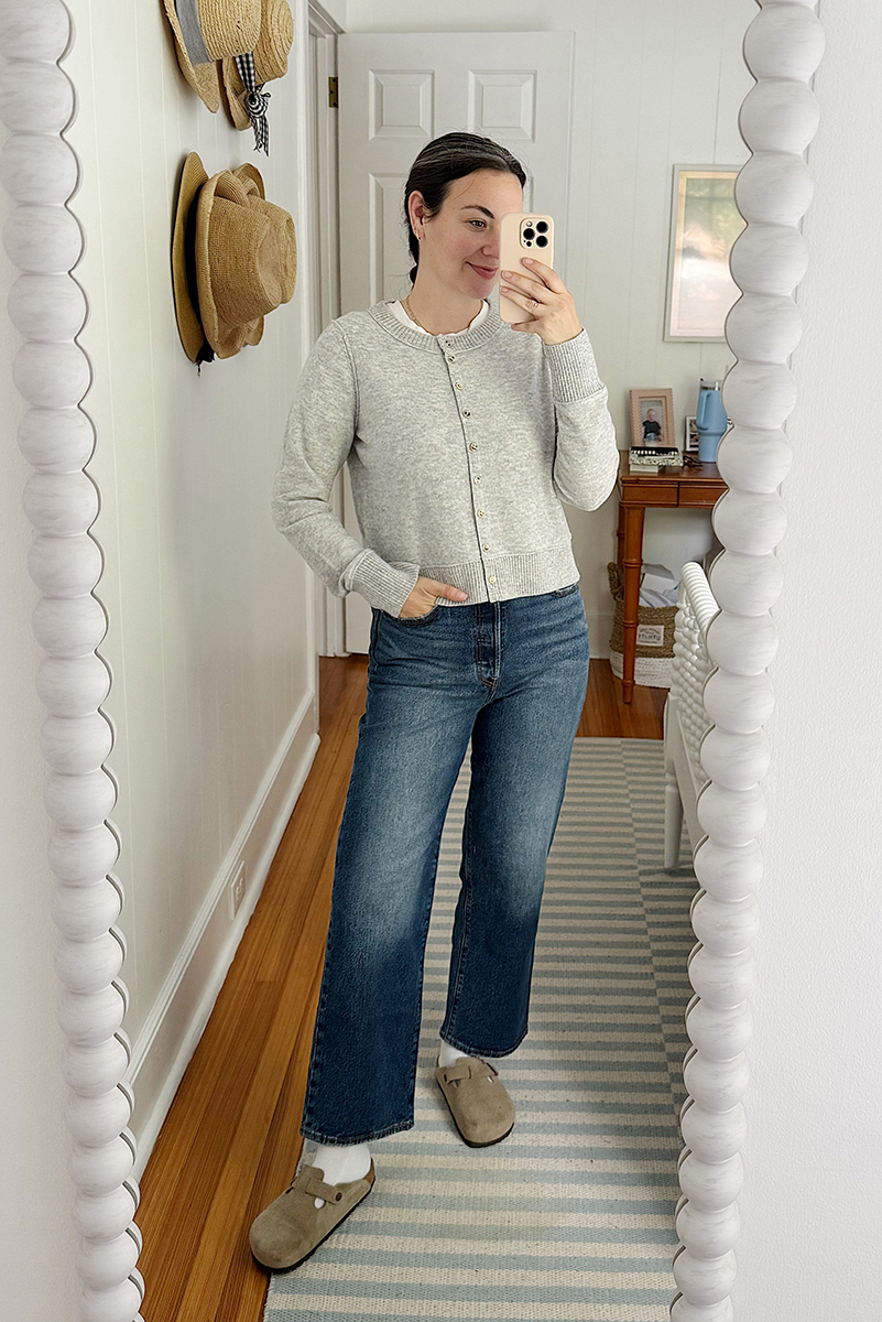 Carly in a mirror selfie wearing a white tee, grey sweater, and wide legged jeans. 