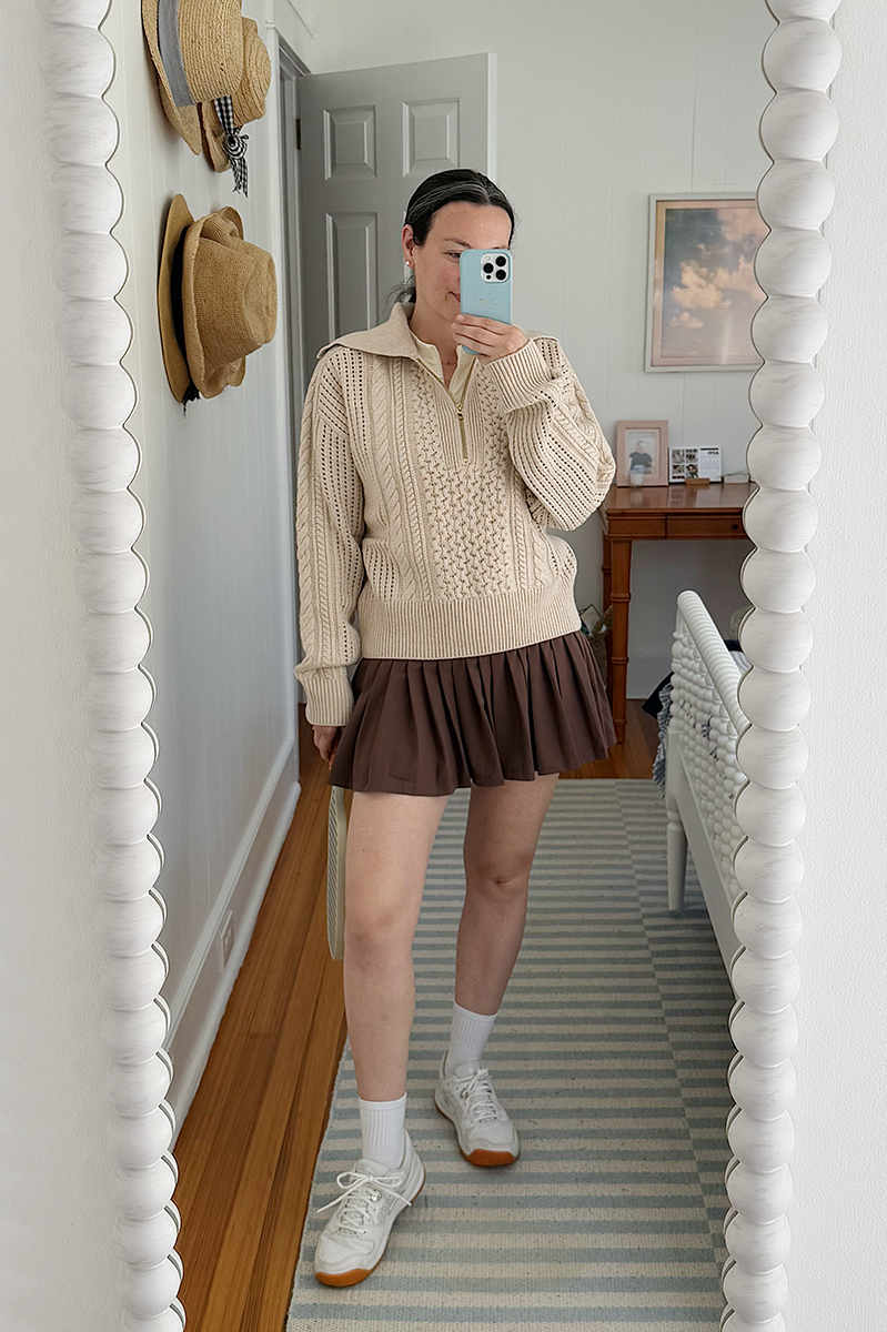 Carly Riordan taking a mirror selfie wearing a beige sweater and brown tennis skirt. 