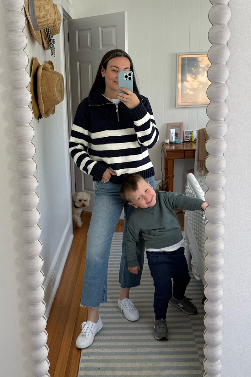 Carly Riordan taking a mirror selfie wearing a navy and white striped pullover, jeans, and sneakers.