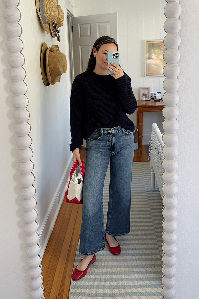 Carly Riordan taking a mirror selfie wearing a navy sweater, wide legged jeans, and red ballet flats