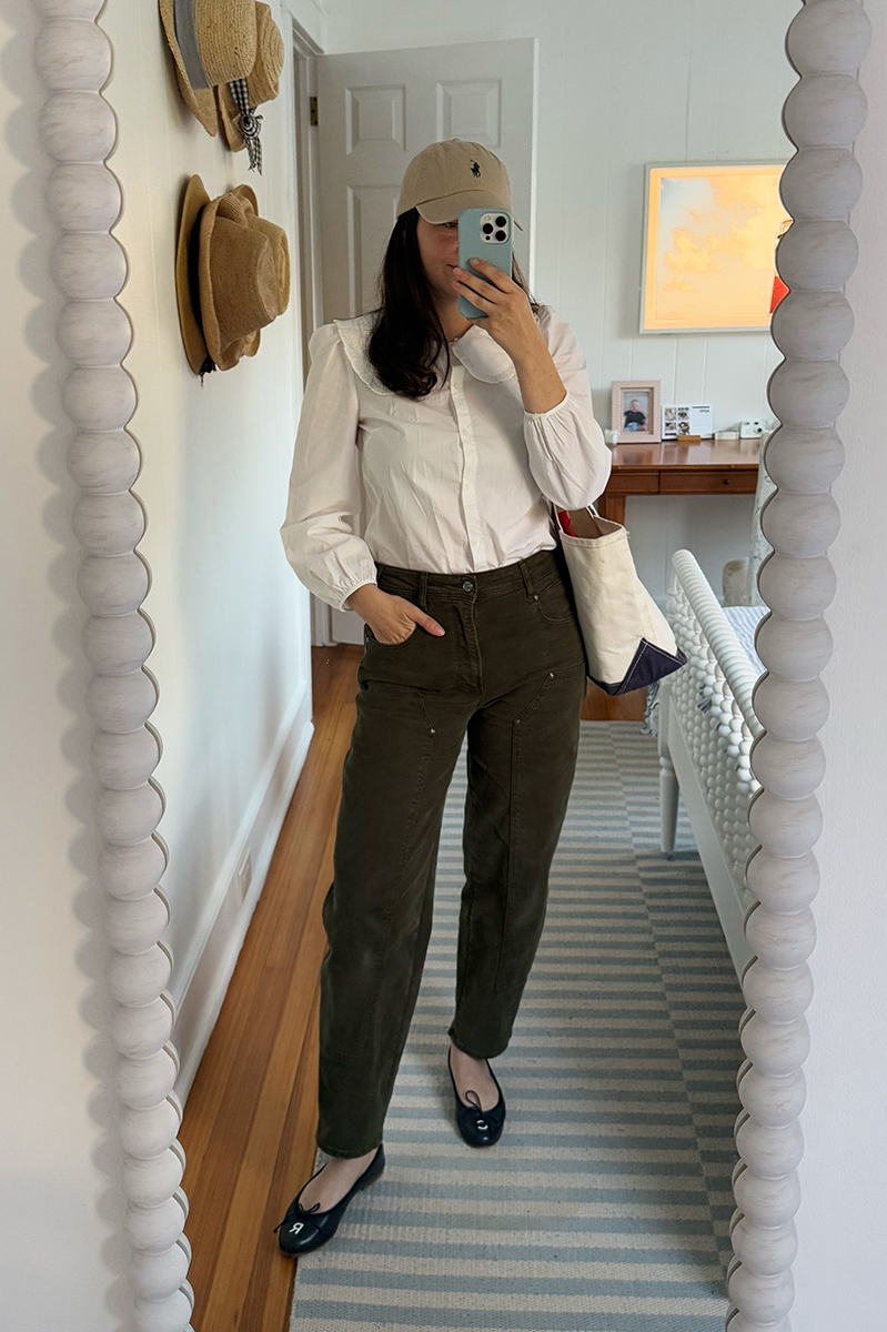 Carly Riordan taking a mirror selfie wearing a white button down, green pants, and navy ballet flats.