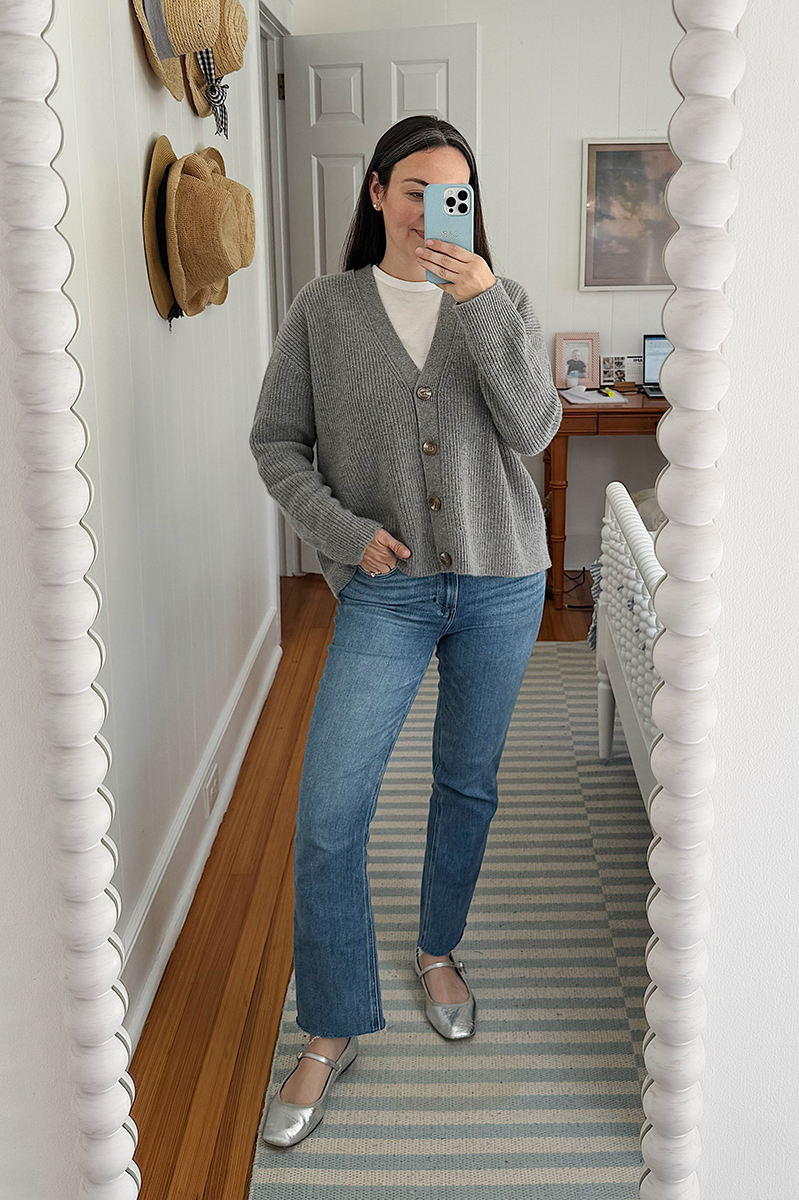 Carly Riordan taking a mirror selfie wearing a grey oversized cardigan, light washed jeans, and metallic ballet flats.