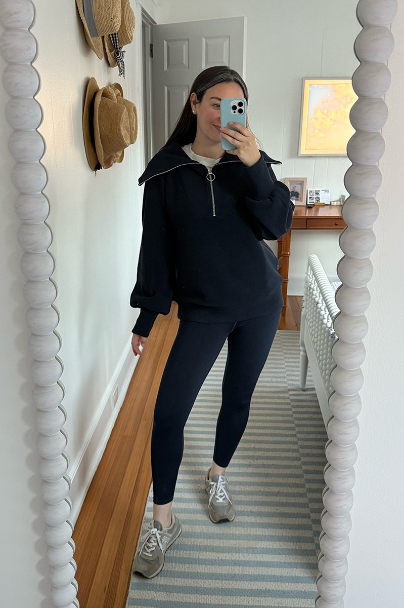 Carly Riordan taking a mirror selfie wearing a navy pullover, navy leggings, and sneakers.