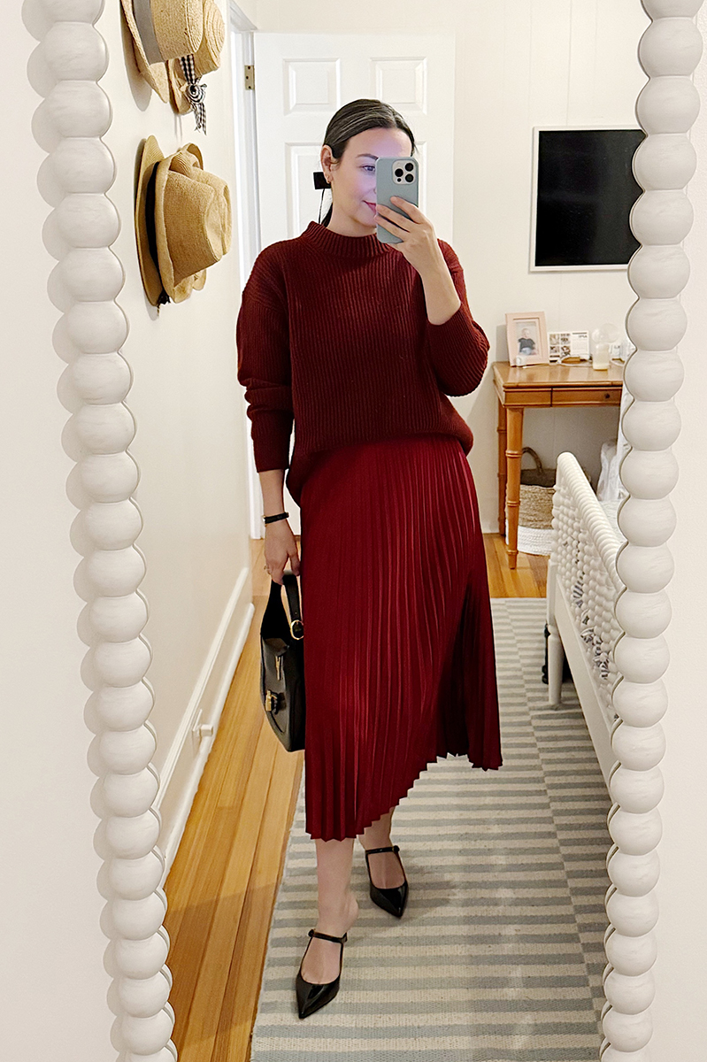 Carly Riordan taking a mirror selfie wearing a pleated silk skirt and cashmere sweater.