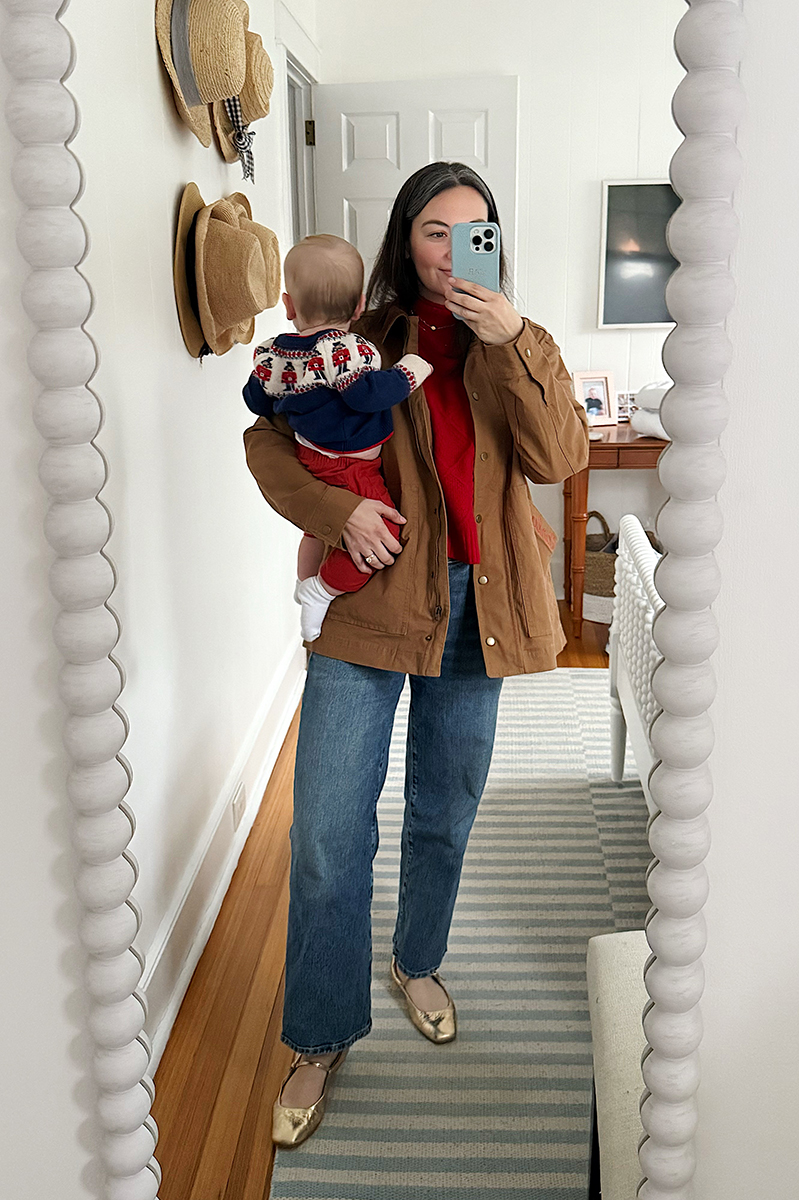 Carly Riordan taking a mirror selfie wearing a barn coat, red sweater and wide legged jeans.