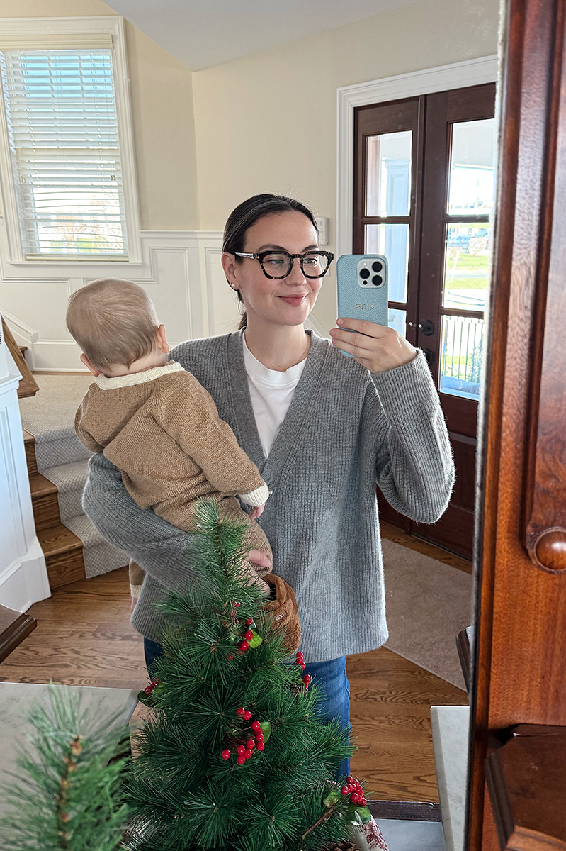 Carly Riordan taking a mirror selfie wearing a grey sweater and glasses.