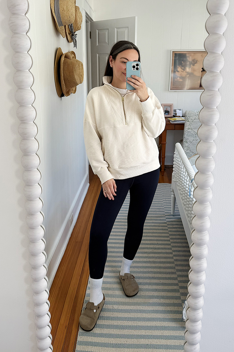 Carly Riordan taking a mirror selfie wearing a cream pullover and birkenstock clogs.