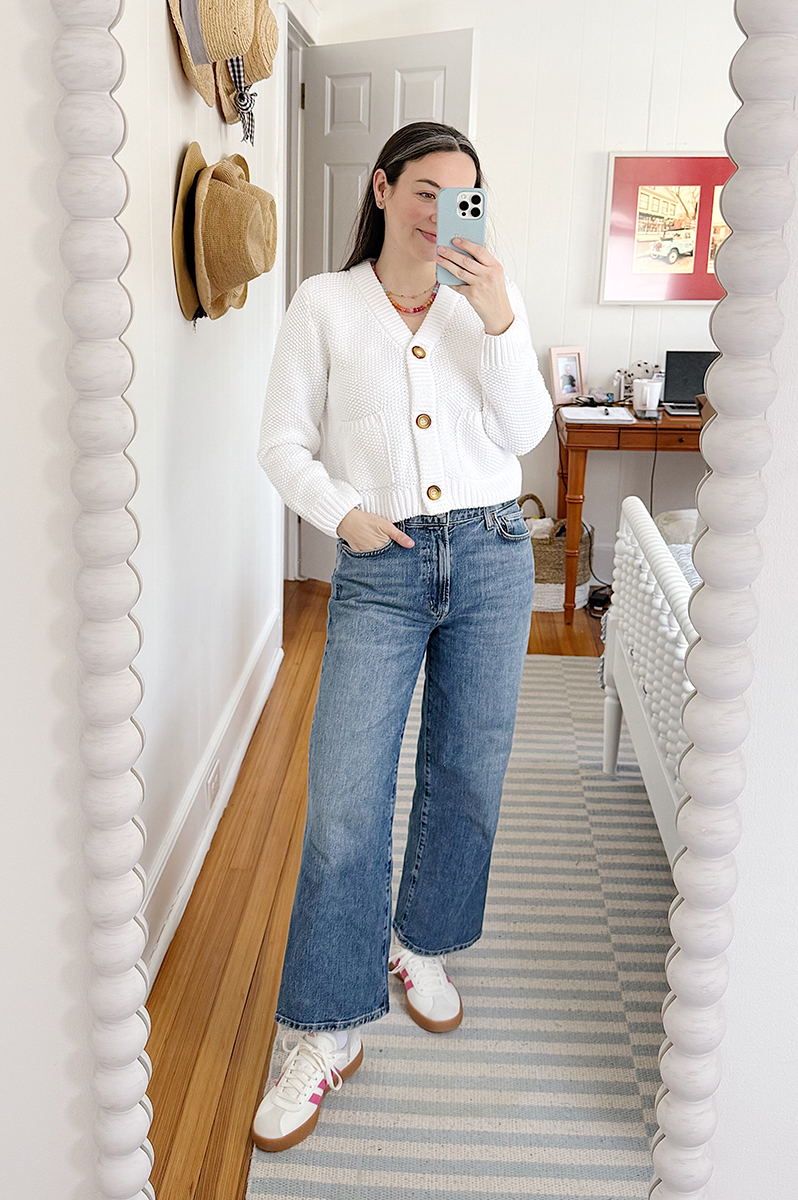 Carly Riordan taking a mirror selfie wearing a white cardigan, jeans, and sneakers.