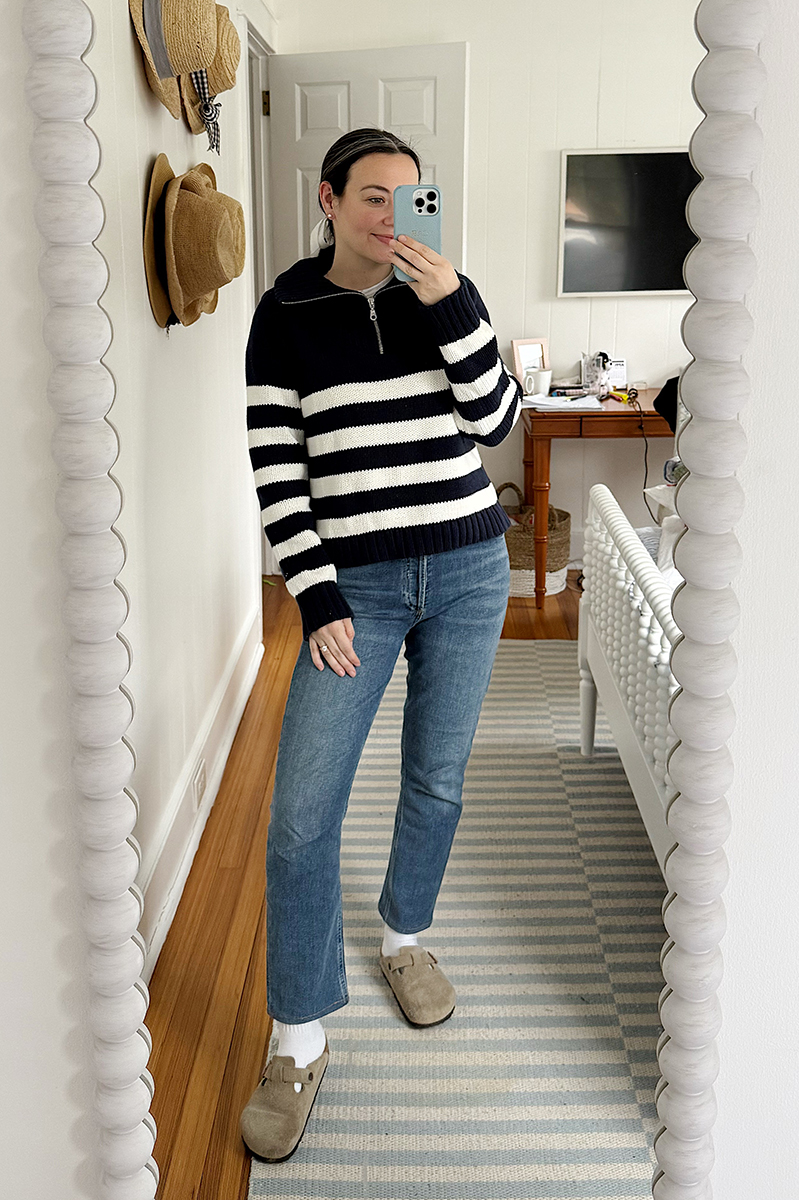 Carly Riordan taking a mirror selfie wearing a navy and white striped sweater and clogs.