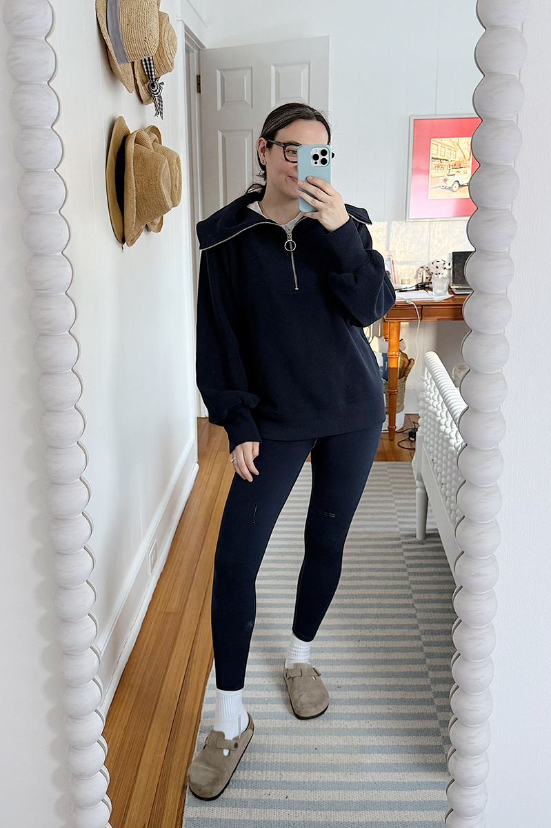 Carly Riordan taking a mirror selfie wearing a navy pullover, leggings, and clogs.