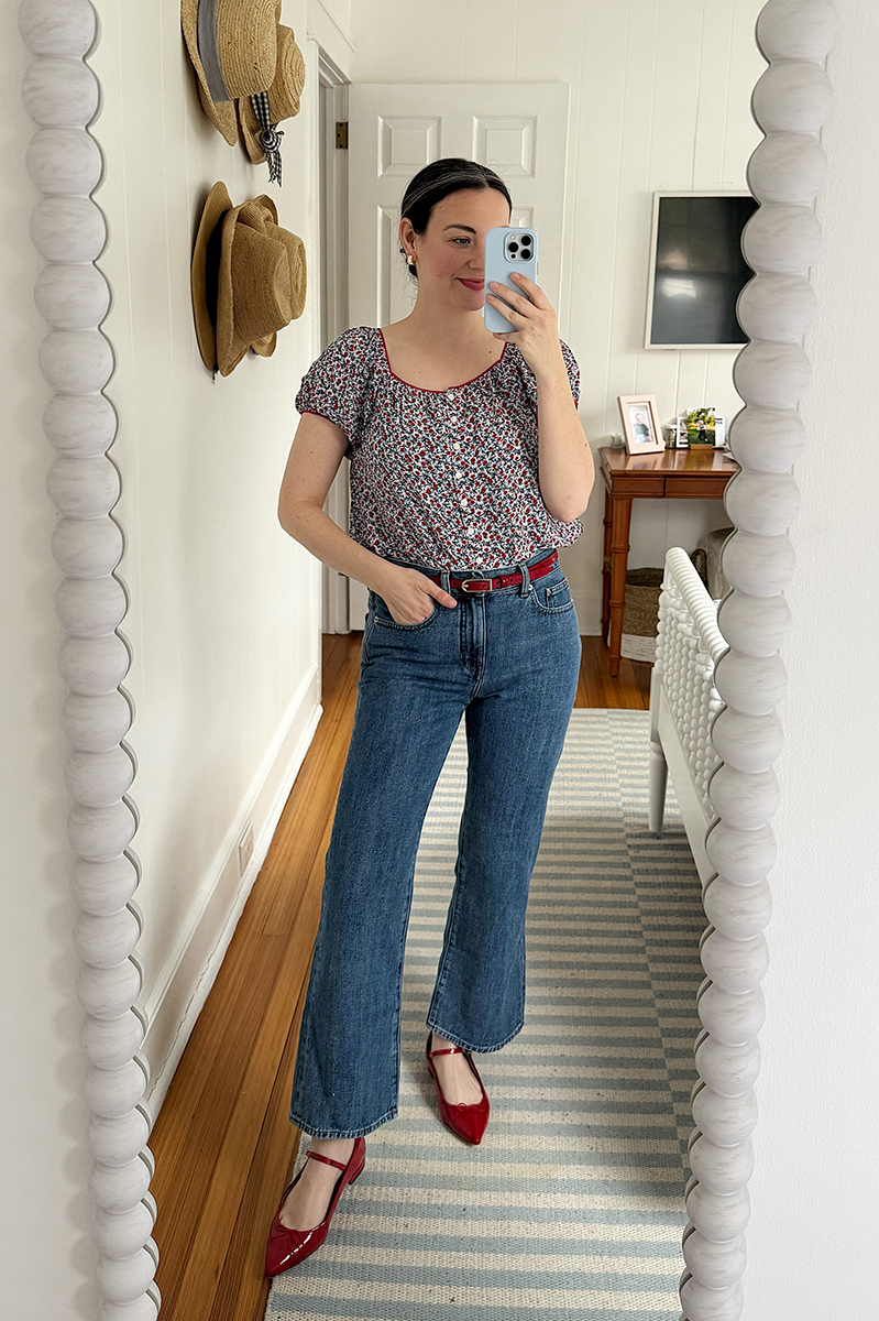 Carly Riordan taking a mirror selfie wearing a floral shirt and red flats.